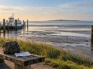 Kleine weiß-blaue Personenfähre liegt bei Ebbe an einem hölzernen Anleger hinter dem Deich, davor weites, feucht glänzendes Watt mit Prielen, Austernfischern und einem Pfahl, am Horizont eine flache Insel; im Vordergrund auf einer verwitterten Holzbank ein Rucksack und ein aufgeschlagener Gezeitenkalender, tiefstehende Morgensonne