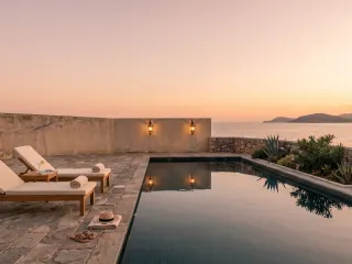 Steinerne Poolterrasse bei Sonnenuntergang, zwei leere Liegen, im ruhigen Wasser spiegeln sich zwei Laternen, dahinter das Meer.