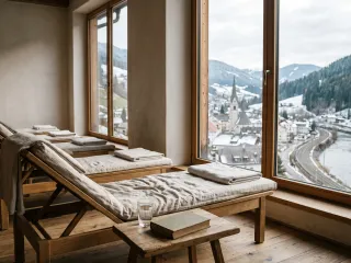 Ruheraum eines kleinen Wellnesshotels mit einer Reihe hölzerner Liegen und gefalteten Leinentüchern vor großen Fenstern, auf einem Holzhocker ein Glas Wasser und ein Buch, draußen ein verschneites Alpental mit Kirchturm, Bahnlinie und Fluss