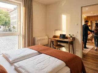 Hotelzimmer mit Doppelbett und Schreibtisch, Fenster zur Altstadtgasse, im Hintergrund ein Paar mit Gepäck vor dem Aufzug