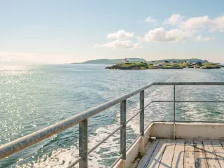 Blick vom Heck einer Fähre über die Reling auf das Kielwasser, dahinter im sonnigen Dunst eine flache grüne Insel mit weißem Leuchtturm und einigen hellen Häusern am Ufer