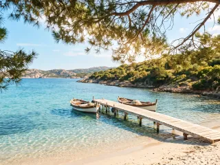 Blick unter Pinienzweigen hindurch auf eine sonnige Mittelmeerbucht: klares türkisfarbenes Wasser, ein hölzerner Steg führt zu zwei kleinen weißen Fischerbooten, rechts ein heller Sandstrand und bewachsene Felsküste