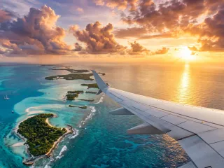 Blick aus dem Flugzeugfenster über die Tragfläche auf türkisfarbene Lagunen und flache Inseln, am Horizont geht die Sonne zwischen Quellwolken unter.