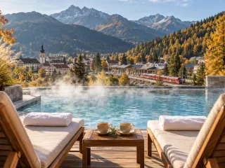 Zwei leere Holzliegen mit gefalteten weißen Handtüchern und zwei Tassen Tee auf einem kleinen Tisch am Rand eines dampfenden Thermalbeckens, dahinter ein kleiner Kurort mit Kirchturm und Bahnhof, in den gerade ein roter Regionalzug einfährt, umgeben von goldgelben Lärchen und verschneiten Berggipfeln im Herbstlicht